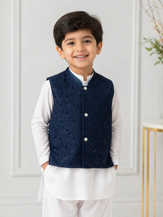 Young boy wearing a navy blue embroidered Waistcoat over a white Shalwar and Kameez, standing against a light-colored wall.